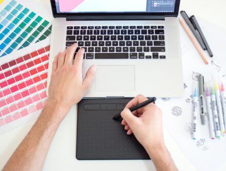 Graphic designer working on a MacBook with colour charts and markers beside the laptop