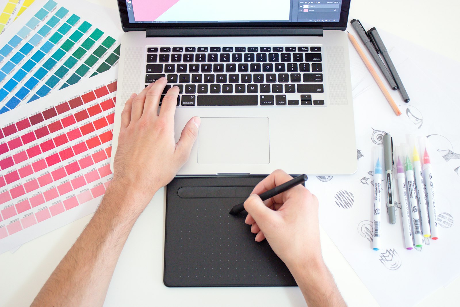 Graphic designer working on a MacBook with colour charts and markers beside the laptop