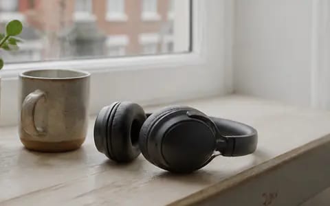A pair of black over-ear headphones rests on a pale wooden windowsill beside a ceramic mug and a potted plant